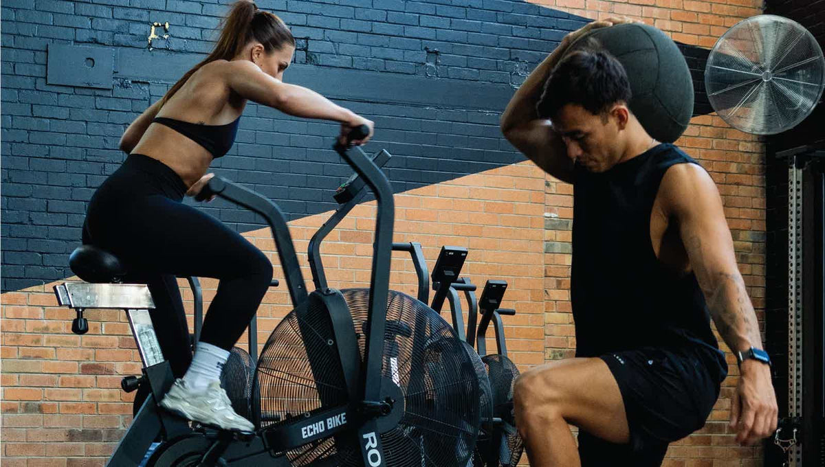 Two people exercising in a gym, one on an exercise bike and one lifting.
