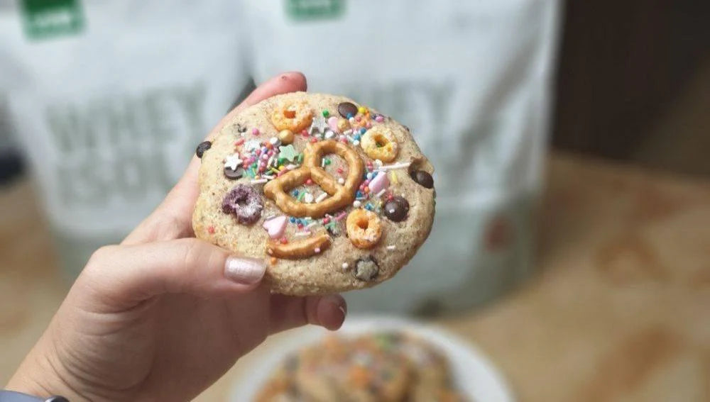 A hand holding a decorated cookie with protein powder bags in the background.