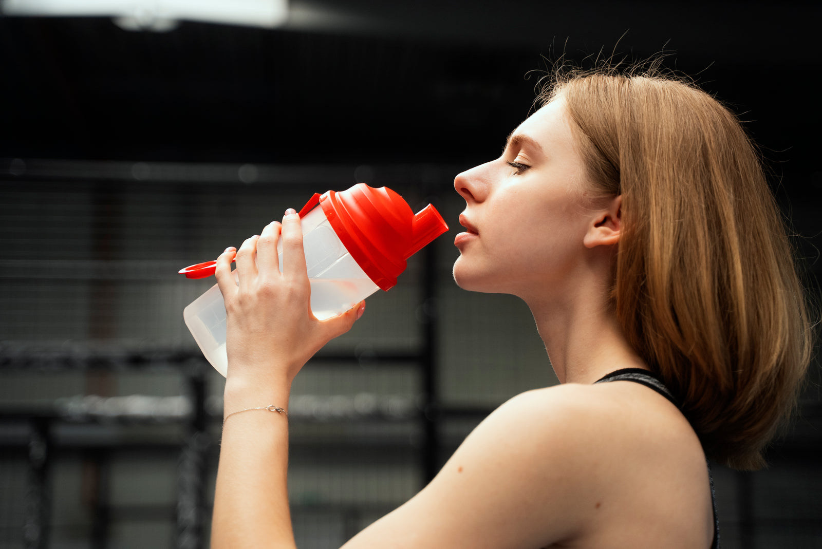 Woman drinking from a red shaker bottle in a gym setting.