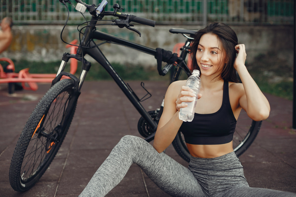 A woman sitting by a bicycle, holding a water bottle, smiling.